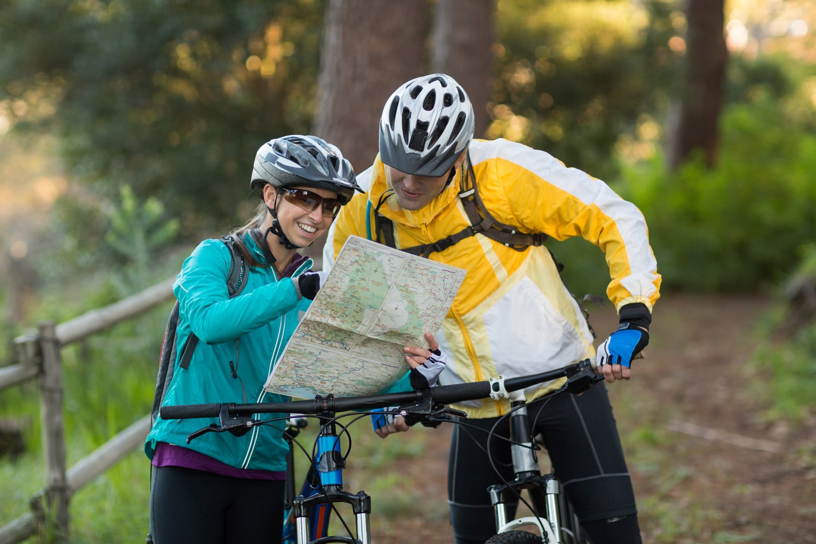 Dois ciclistas de capacete em pé com suas bikes em uma trilha na floresta, sorrindo enquanto consultam juntos um mapa de papel
